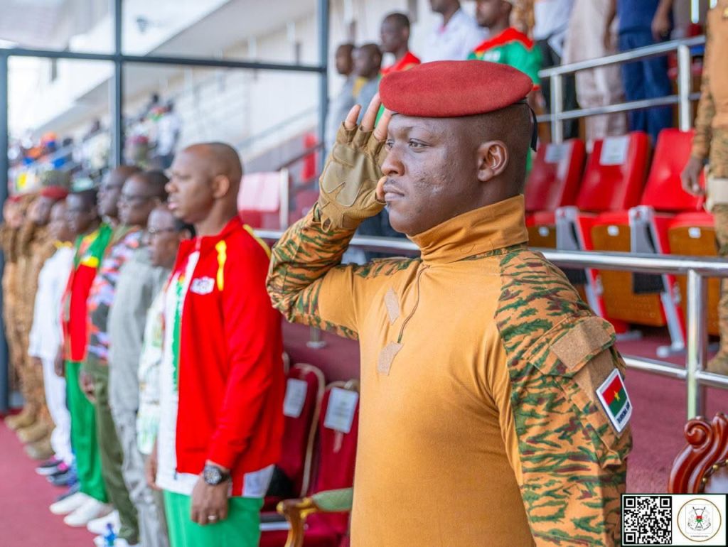 Un soldat en uniforme militaire saluant, avec des collÚgues en arriÚre-plan, lors d'une cérémonie officielle.
